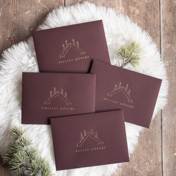 Vier Umschläge in der Farbe burgunder, alle haben eine Prägung mit einem Schwibbogen und 1-4 leuchtende Kerzen und den Worten "erster Advent", "zweiter Advent" etc.